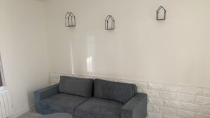 Un canapé gris avec deux coussins, une petite table ronde aux pieds en bois et un mur blanc décoré d’un motif imitant des briques.