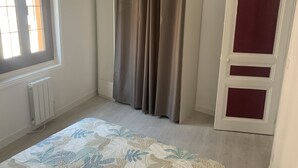 Une chambre avec une fenêtre, un radiateur, un lit recouvert d’une couvre-lit à motifs floraux, une armoire et une porte avec un panneau rouge.