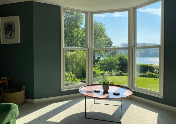 A room with a large bay window offering a view of a lush garden and a body of water. There is a round table with a potted plant in the center.