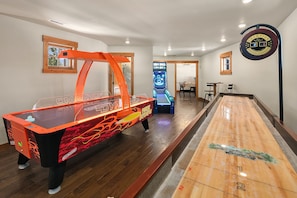 Una mesa de shuffleboard, una mesa de fútbolín y una mesa de billar en una habitación con piso de madera y paredes blancas.