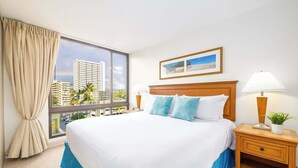 A hotel room with a large bed, a window with a view of buildings, a framed picture on the wall, and a bedside table with a lamp and a plant. - 2BR Condo w/ lanai & Diamond Head views (Honolulu)