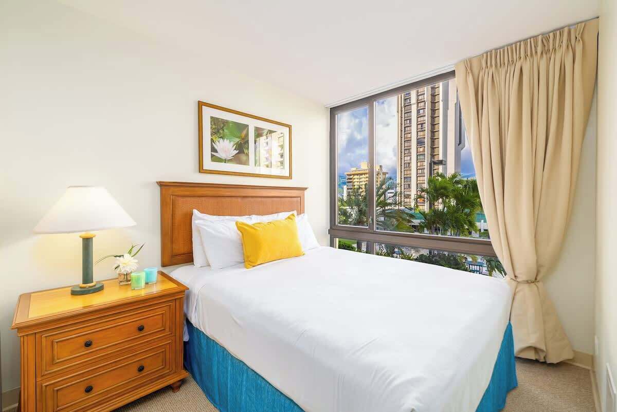 Habitación de hotel con cama, mesita de noche, lámpara, un cuadro enmarcado y un ventanal con cortinas.