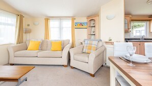 Een gezellige woonkamer met een bank en fauteuil, een houten salontafel, en een keukenhoek met een eettafel gedekt voor twee personen.