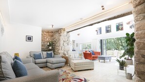 Ein geräumiges Wohnzimmer mit Steinwand, einem grauen Sofa, einem roten Sessel und einem großen Fenster mit Blick auf einen Garten.