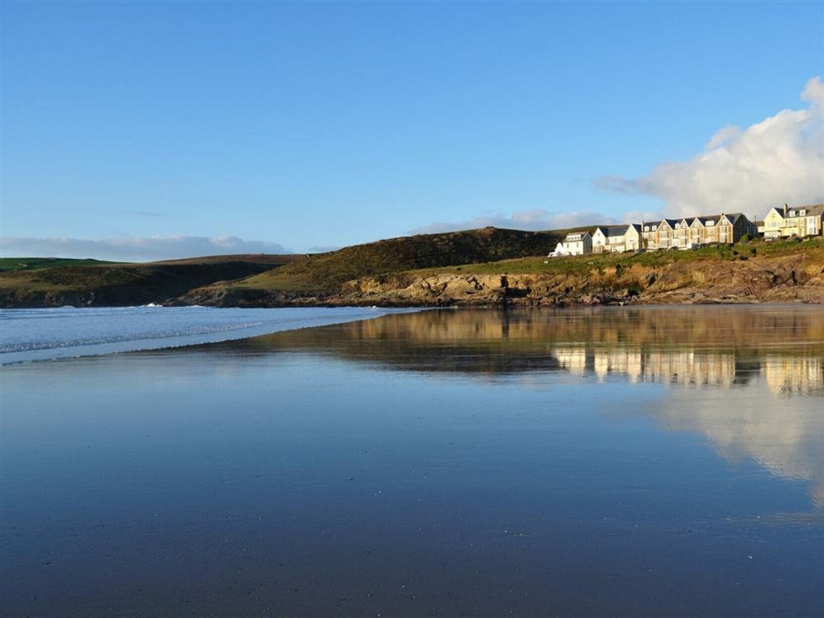 A coastal scene with a beach, calm water, and a row of white houses with gabled roofs on a hillside.