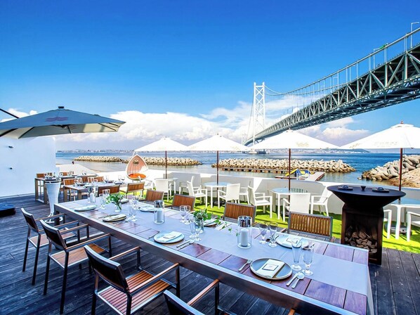 An outdoor dining area with tables set for a meal, overlooking a body of water and a bridge.