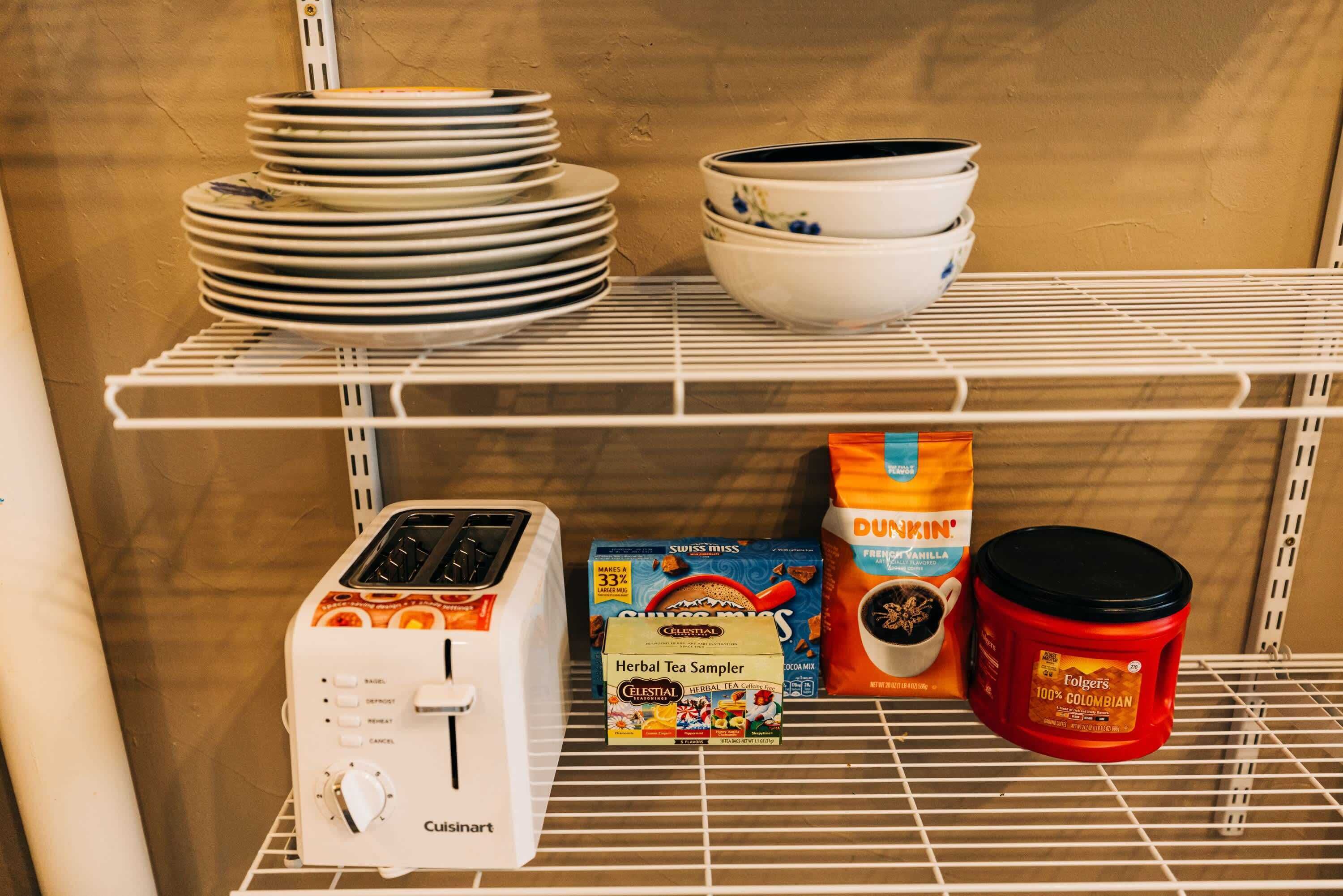 A kitchen shelf with stacked plates, bowls, a toaster, a box of herbal tea sampler, a bag of Dunkin' coffee, and a red mug.