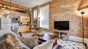 Ein modernes Wohnzimmer mit einer Ledersofa, einem Couchtisch und einem Fernseher. Im Hintergrund ist eine Ziegelwand und eine Küche erkennbar.