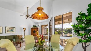 Un salon moderne avec une table en verre, des fauteuils verts, un ventilateur de plafond et une grande fenêtre donnant sur l’extérieur.