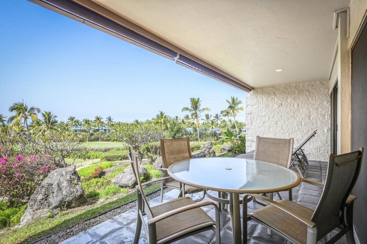 A balcony with a glass table and chairs, overlooking a landscaped garden with palm trees and a view of a residential area.