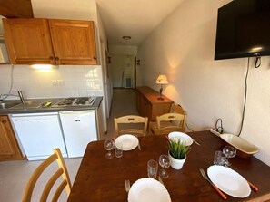 A dining area with a wooden table set for four, a kitchen with white appliances, and a flat-screen TV mounted on the wall.
