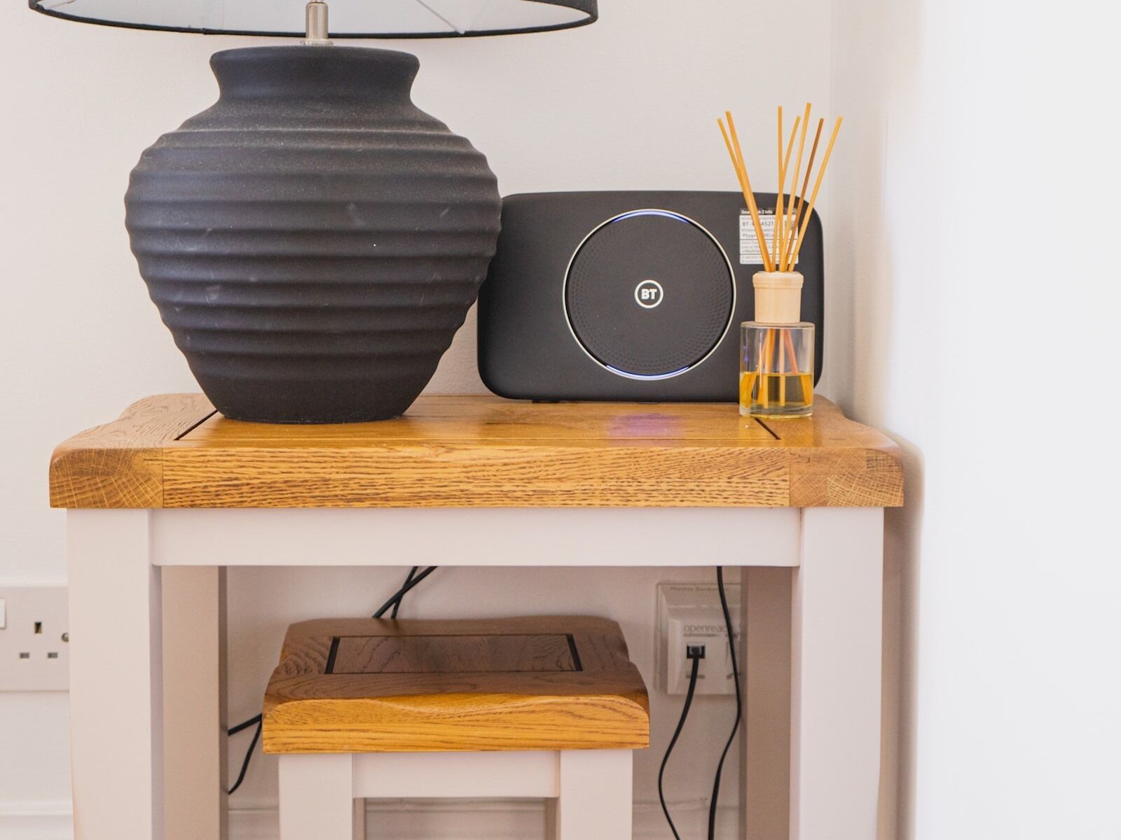 A black speaker with a circular logo, a black vase with a textured surface, and a glass with a wooden stick holder on a wooden shelf.