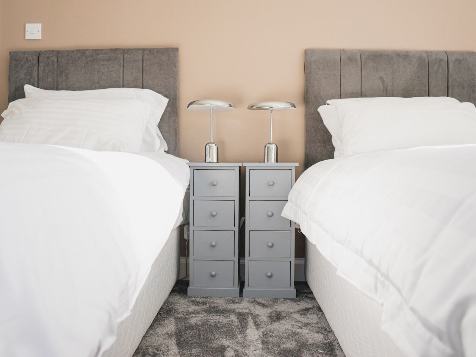 A double room with two beds, grey headboards, white bedding, and a grey nightstand with two lamps.