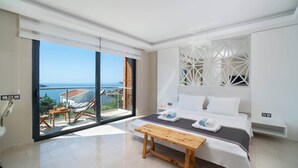 Une chambre moderne avec un grand lit, un banc en bois et une vue sur la mer grâce à de grandes fenêtres du sol au plafond.