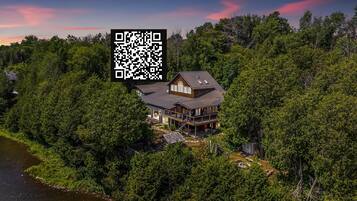 A large wooden cabin with a deck, surrounded by dense forest and a lake.