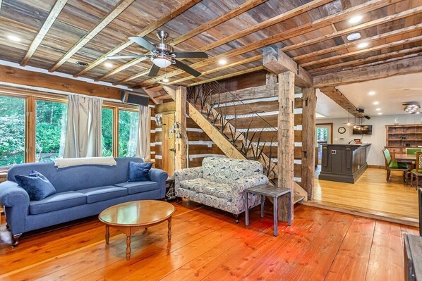 A spacious living room with a wooden ceiling, a blue sofa, a patterned sofa, a coffee table, and a staircase leading to another level.