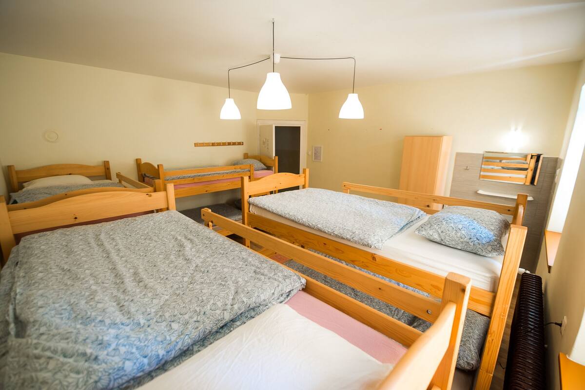 A dormitory room with four wooden bunk beds, each with a pillow and blanket. There are two pendant lights hanging from the ceiling.