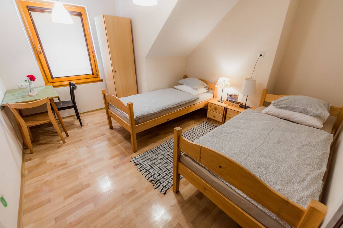 A small, well-lit bedroom with two single beds, a wooden desk, a chair, and a window.