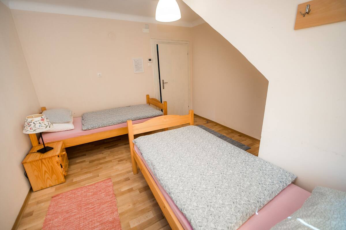 A small, well-lit room with two single beds, a wooden bench, a small table, and a door.