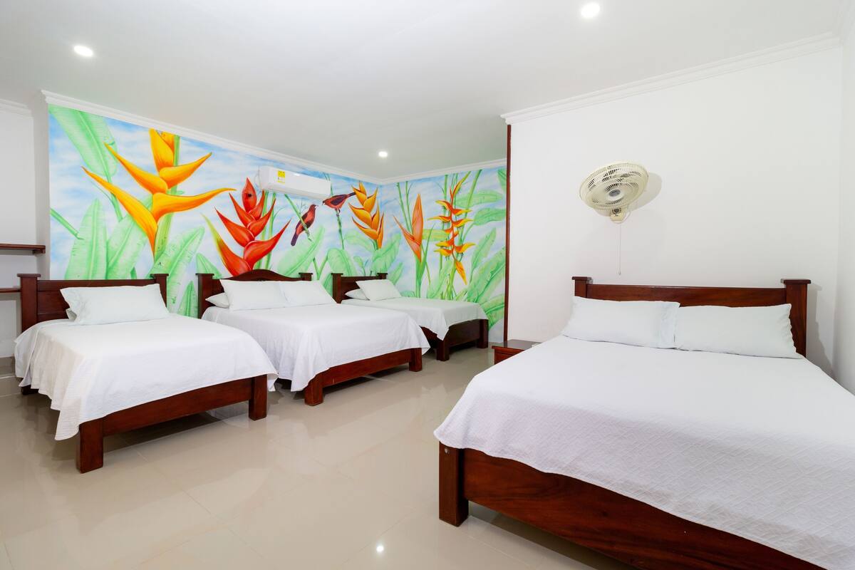 Une chambre d’hôtel avec trois lits en bois, du linge de lit blanc et une fresque représentant des plantes tropicales sur le mur.