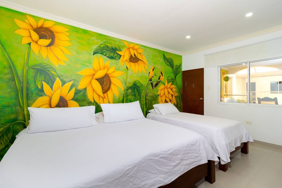 Une chambre d’hôtel avec deux lits, une fresque murale verte représentant des tournesols et une fenêtre donnant sur un paysage agréable.