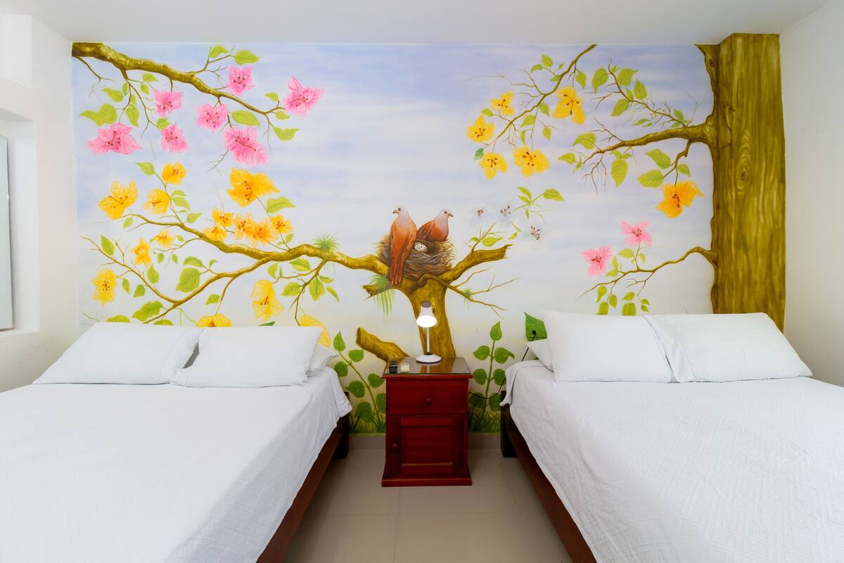 Une chambre avec deux lits, une table de chevet avec une lampe et une fresque représentant un arbre avec des oiseaux et des fleurs.