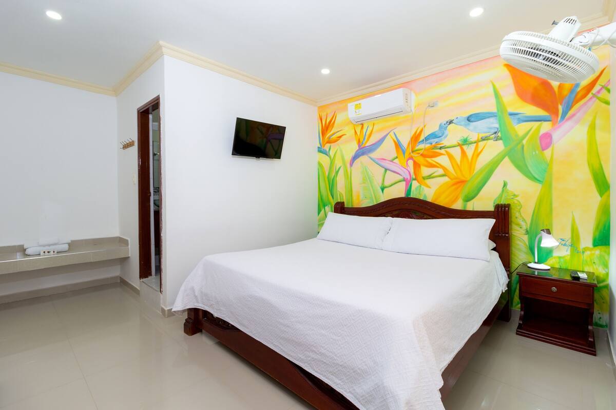 Une chambre d’hôtel équipée d’un lit, d’une table de chevet, d’une télévision, d’un climatiseur et d’une fresque murale aux couleurs vives.