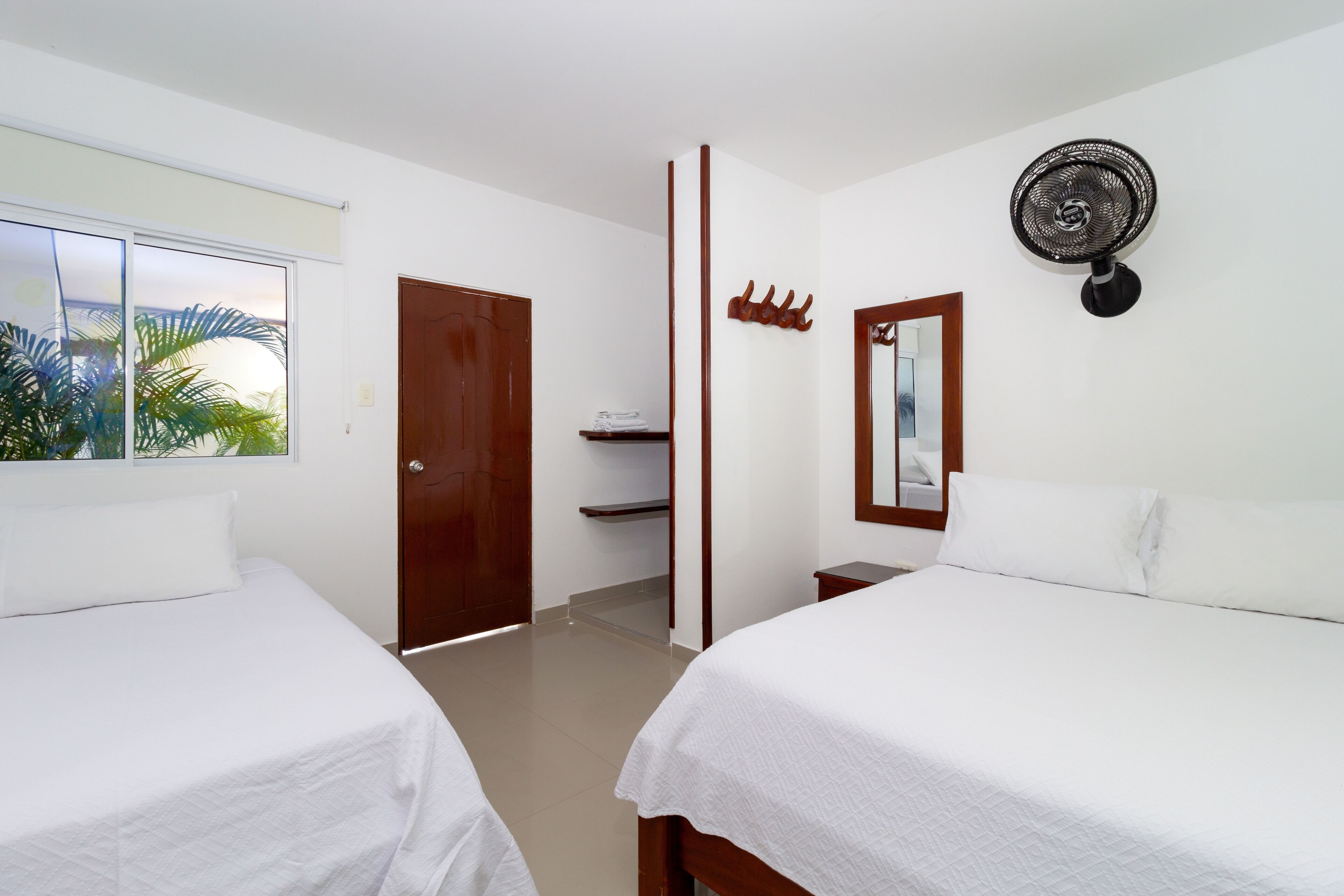 Une chambre d’hôtel avec deux lits, une fenêtre donnant sur des palmiers, une porte en bois, un ventilateur fixé au mur et une petite étagère avec un miroir et des crochets.