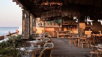 An outdoor dining area with wooden tables and chairs, a stone wall, and a view of the sea.