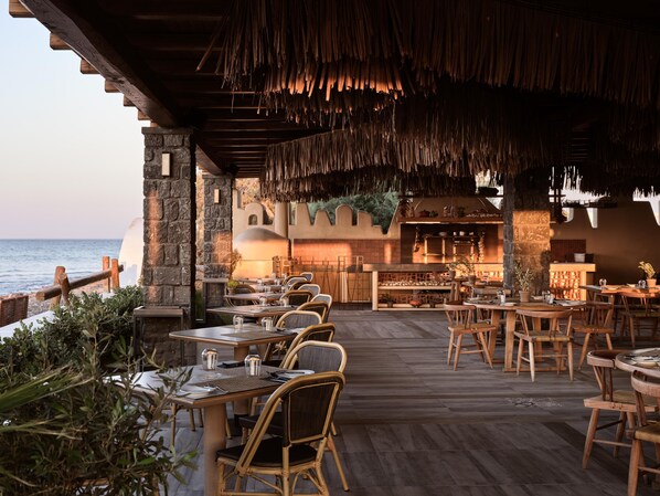 An outdoor dining area with wooden tables and chairs, a stone wall, and a view of the sea.