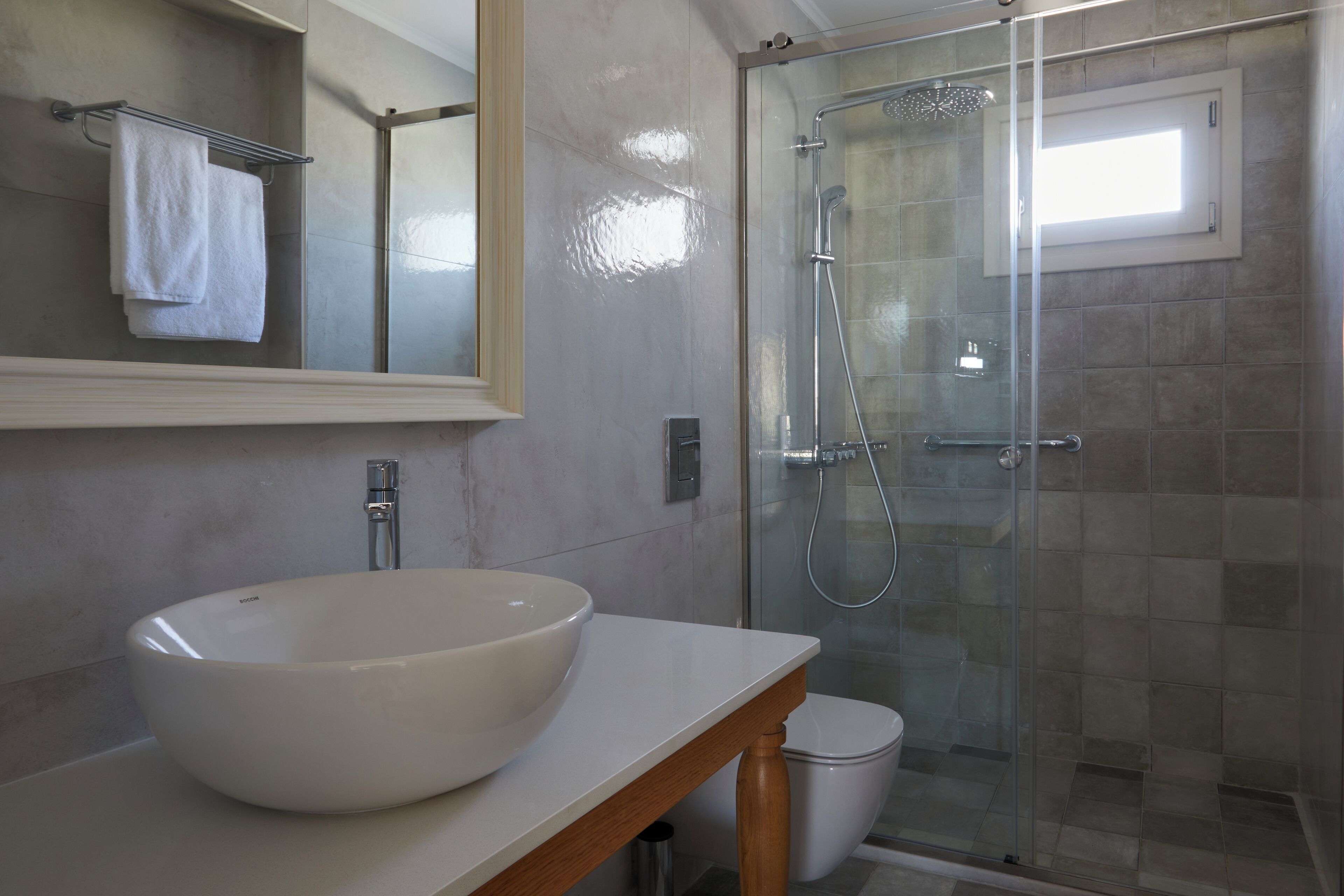 A modern bathroom with a large white vessel sink, a glass-enclosed shower, and a small window above the toilet.