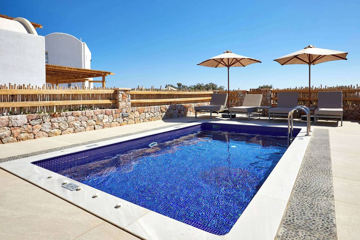 Une piscine rectangulaire bordée de carreaux, entourée de chaises longues et de parasols, adossée à un mur de pierre et se fondant dans un ciel bleu azur.