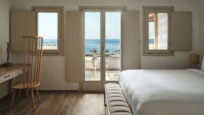 Een slaapkamer met een groot bed, een houten stoel en een bureau. Drie ramen bieden uitzicht op het strand en de oceaan.