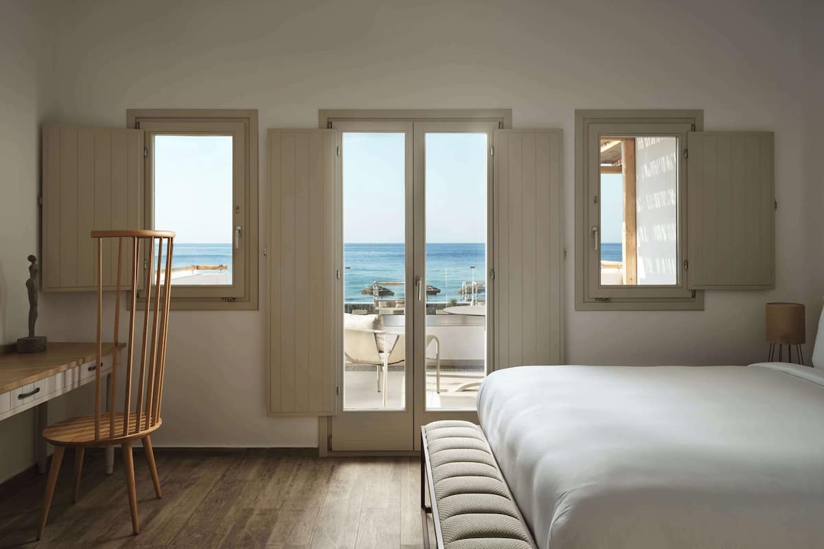 Une chambre à coucher avec un grand lit, une chaise en bois et un bureau. Trois fenêtres offrent une vue sur la plage et l’océan.
