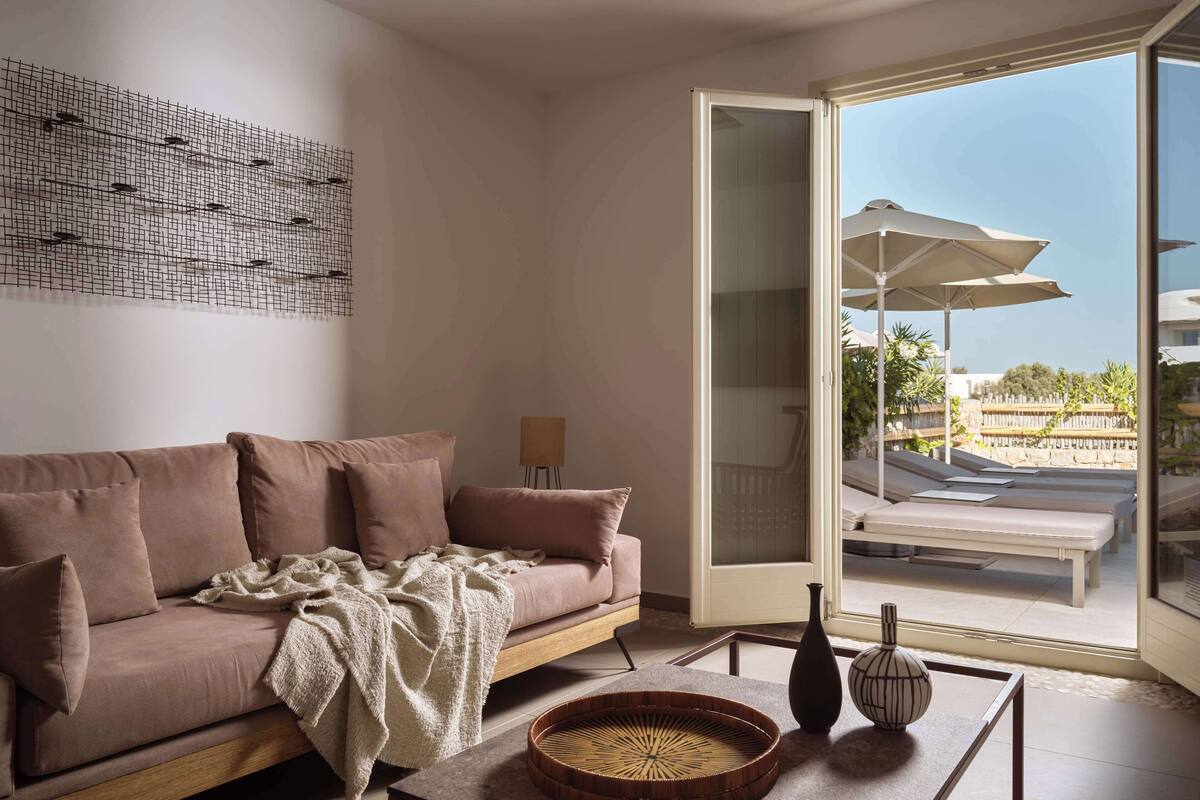 Un salon moderne comprenant un canapé, une table basse agrémentée d’objets décoratifs, un tableau au mur et une porte-fenêtre coulissante donnant sur un espace extérieur équipé de parasols.