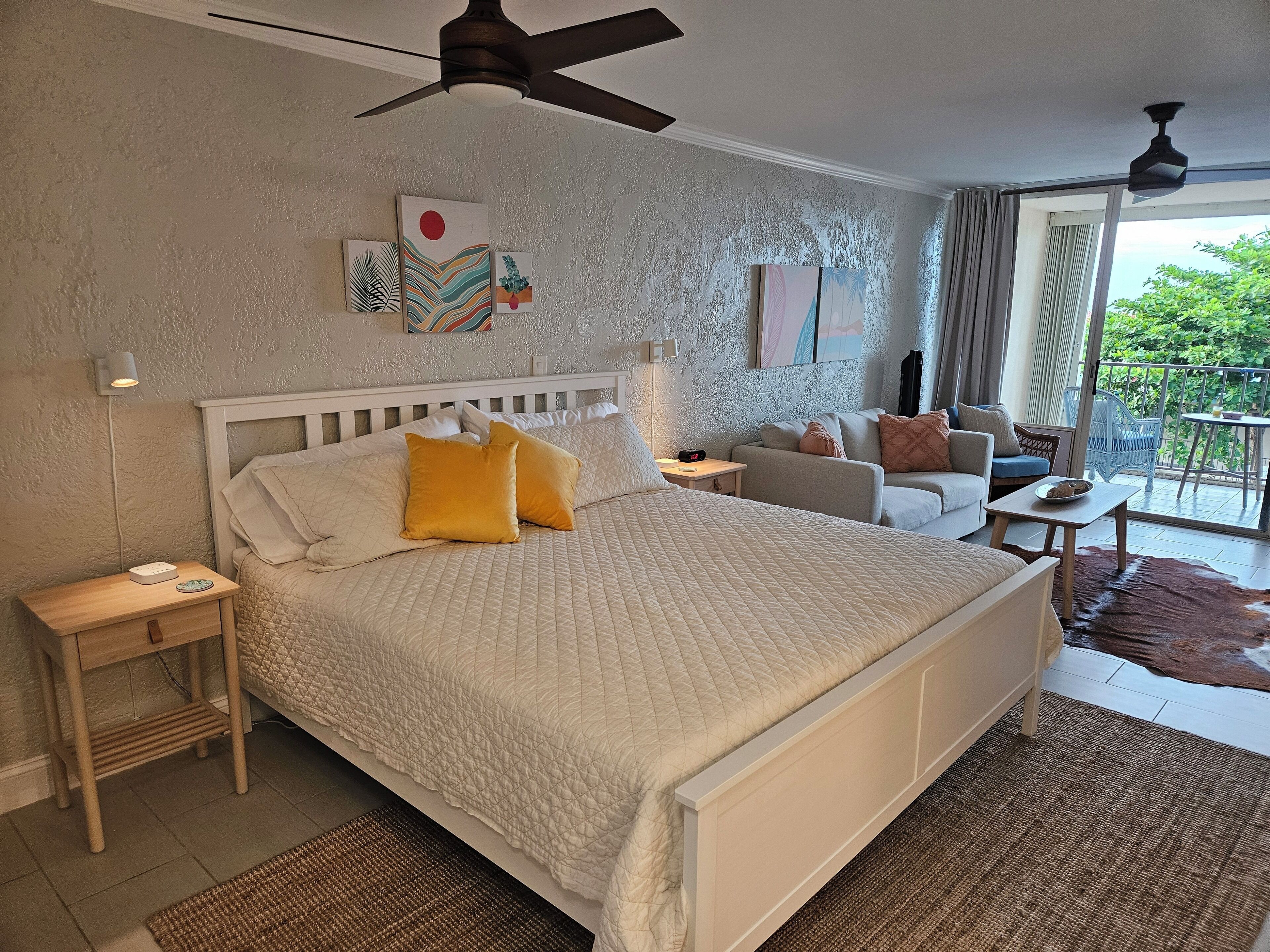 A bedroom with a large bed, a nightstand, a bedside table, a ceiling fan, and a view of the outdoors.