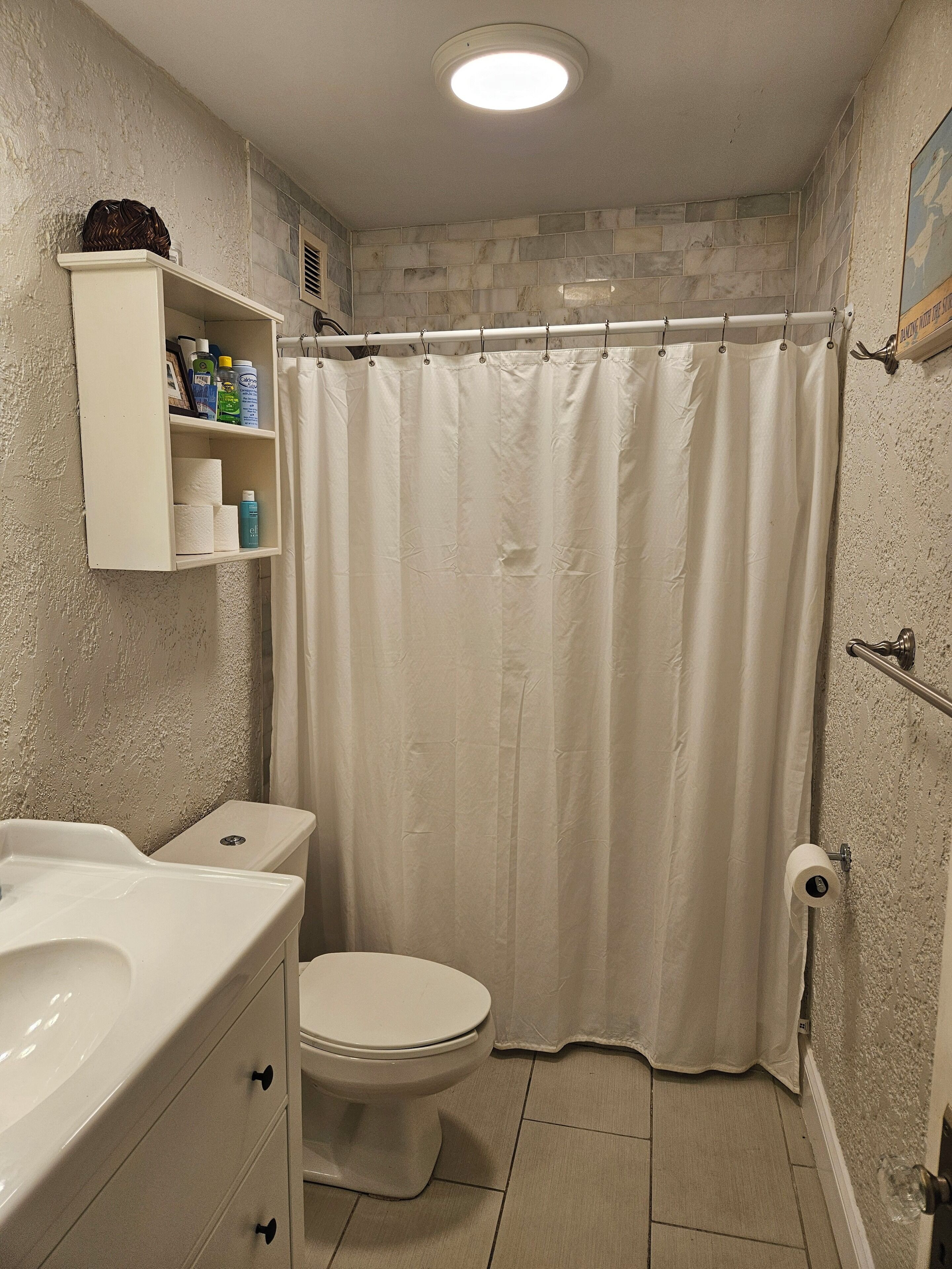 Un baño con inodoro, lavamanos, cortina de ducha y estante con artículos de aseo.
