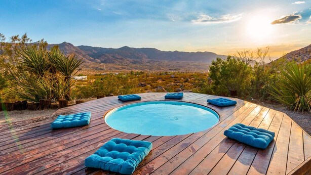 A wooden deck with a circular pool and blue cushions, surrounded by desert vegetation and mountains. - Neon Moon: The Cosmic Experience w/ Pool & Spa (Joshua Tree)