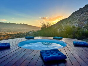 Une terrasse en bois avec un jacuzzi, entouré de coussins bleus, offrant une vue sur un paysage montagneux au coucher du soleil.