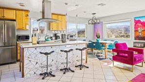 A modern kitchen with a central island, bar stools, and a dining area with chairs and a table. - Neon Moon: The Cosmic Experience w/ Pool & Spa (Joshua Tree)