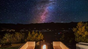 A night sky with visible stars and the Milky Way over a patio area with two cushioned chairs and a fire feature. - Neon Moon: The Cosmic Experience w/ Pool & Spa (Joshua Tree)