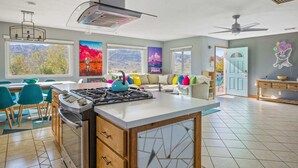 A modern kitchen with a gas stove, dining area with chairs, and large windows with mountain views. - Neon Moon: The Cosmic Experience w/ Pool & Spa (Joshua Tree)