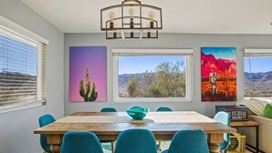 A dining area with a wooden table, teal chairs, and a central teal bowl. There are three wall-mounted paintings depicting cacti and landscapes. - Neon Moon: The Cosmic Experience w/ Pool & Spa (Joshua Tree)