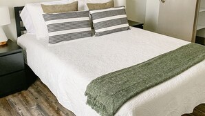 A neatly made bed with a white duvet and striped pillows, a green throw blanket, and a painting of a rural landscape above it.