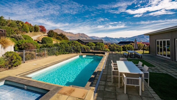 A swimming pool with a view of mountains, a patio with outdoor furniture, and a fenced garden area.