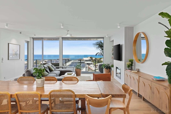 A modern living room with a large dining table, comfortable seating, a TV, and a view of the ocean.