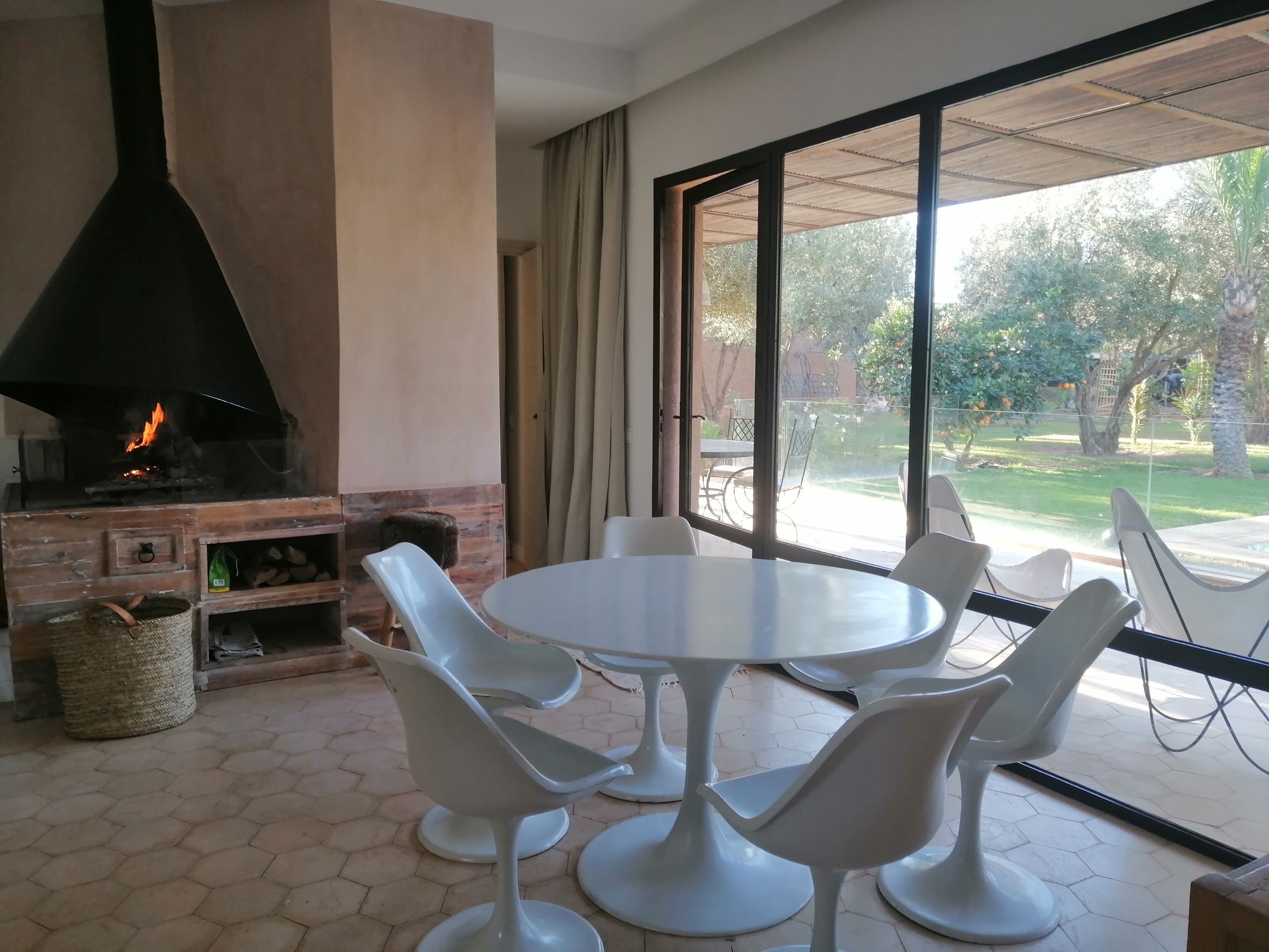 Een eetruimte met een ronde tafel en witte stoelen, een open haard van baksteen en uitzicht op een tuin door grote ramen.