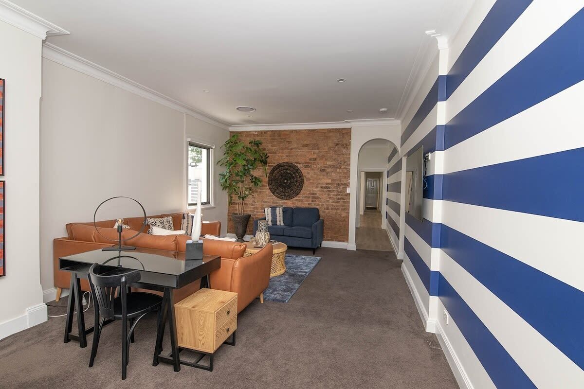 Una sala de estar moderna con pared de ladrillo decorativa, pared azul y blanca a rayas, sofá de cuero marrón, sofá azul, mesa de comedor con sillas y planta en maceta.