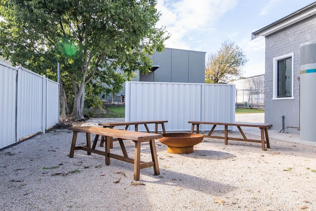 An outdoor patio area with three wooden picnic tables and a fire pit, surrounded by a gravel surface and metal fencing.