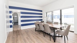 A modern dining area with a long table and six chairs, a blue and white striped wall, and a view through large windows. - McNamara Lane - CBD, 7 Smart TVs, Sleeps 11 (Orange)